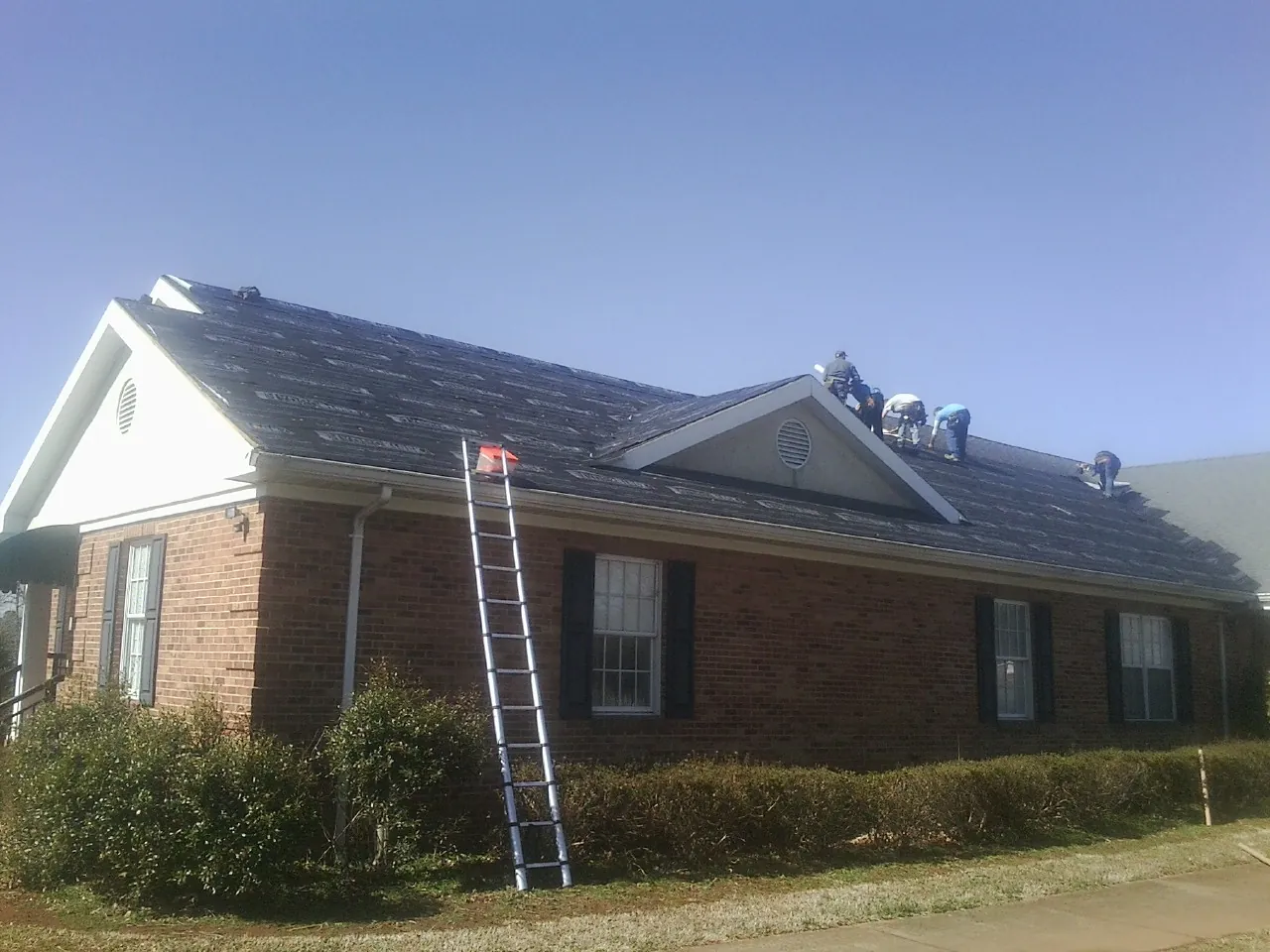 Crew tearing shingles off a brick ranch roof with a ladder set — Emergency Roof Repair work by our crew in Carmine
